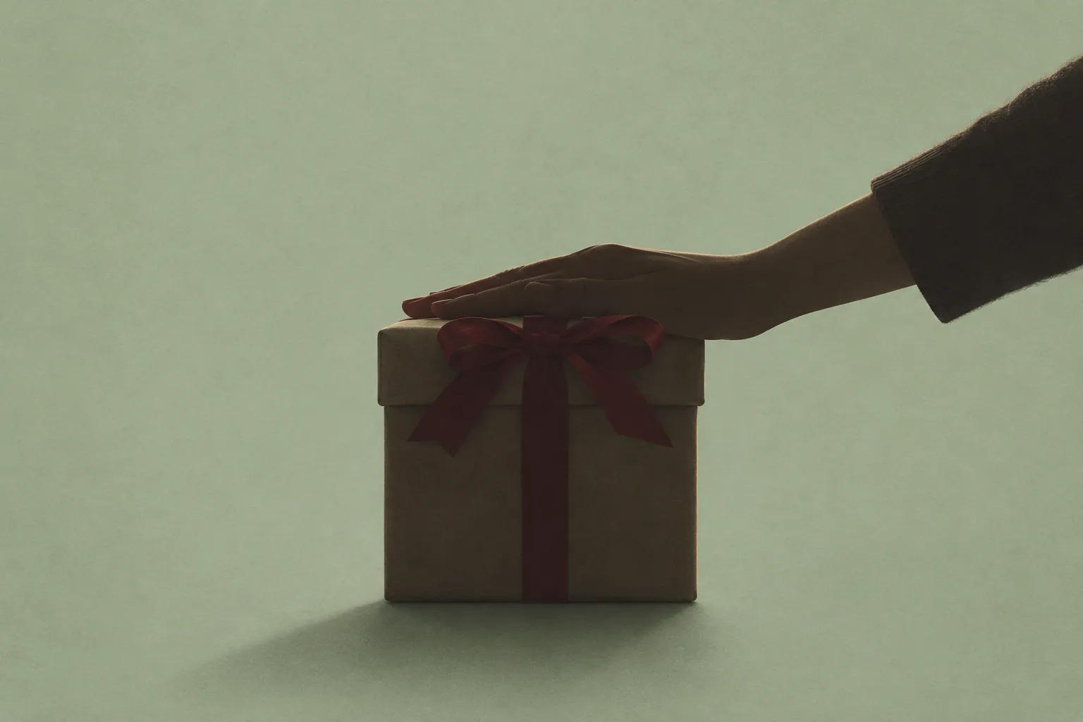 AI-genererad illustration: En hand vilar på locket till ett paket inslaget i brunt papper med ett mörkrött sidenband och rosett, mot en dämpad grågrön bakgrund.